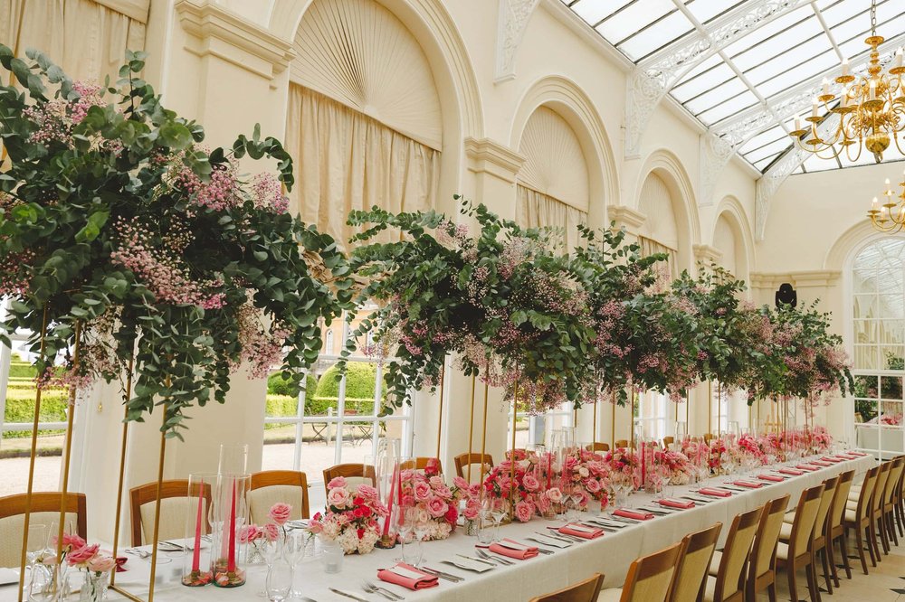 Wedding table setting featuring pink roses, cascading greenery, red candles, and crystal glasses, under a decorated archway in an ornate venue.